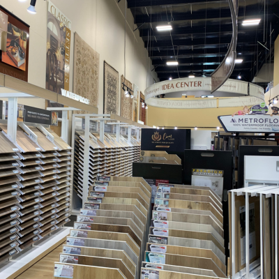 Flooring store aisle with stacked vinyl displays, brochures on stands, and shelves along the wall under bright ceiling lights, with a IDEA CENTER sign above.